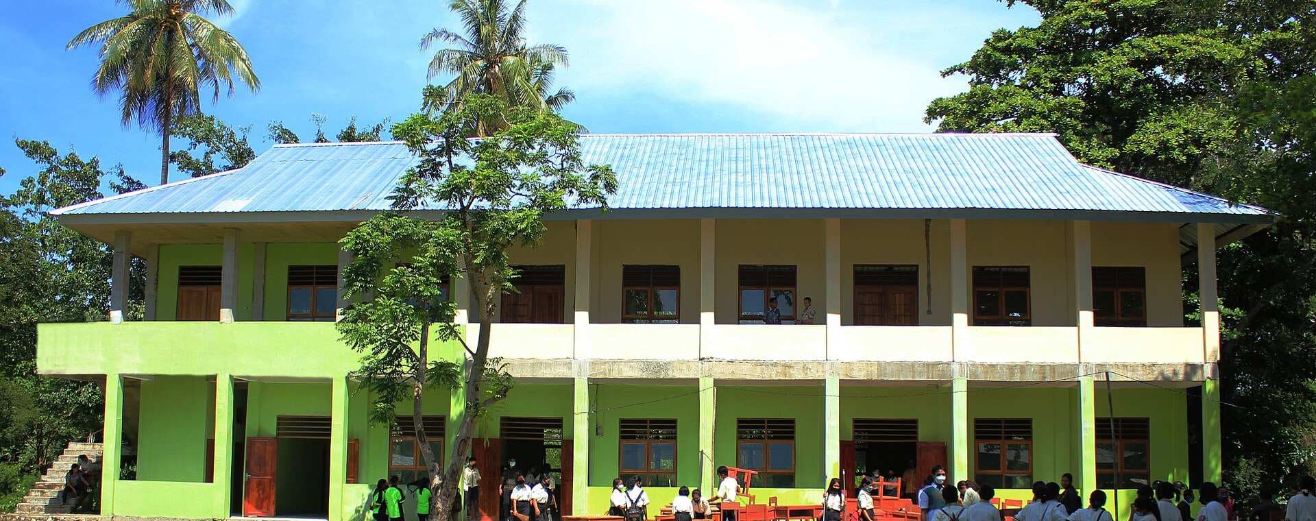 Gedung sekolah menengah dua lantai berwarna hijau dengan siswa berkumpul di koridor lantai dasar